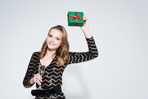 cheerful young woman holding glass of champagne and wrapped christmas present on grey