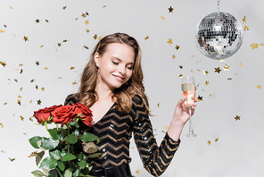 cheerful young woman holding red roses and glass of champagne near disco ball and falling confetti on grey