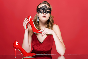 young elegant woman in black carnival mask holding shoe on red