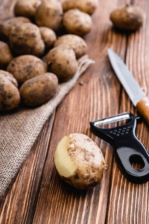 selective focus of dirty peeled potato on wooden table with peeler and knife