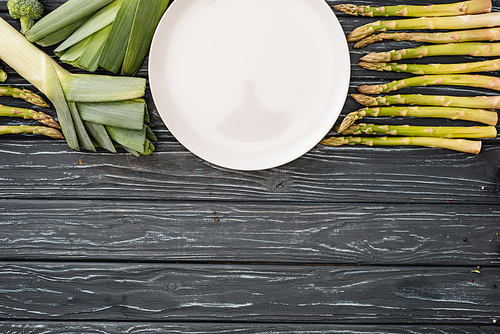 top view of fresh green leek, asparagus and empty plate on wooden surface