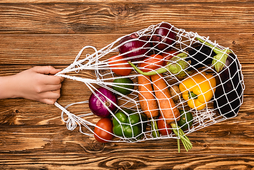 cropped view of woman holding string bag with fresh ripe vegetables on wooden table