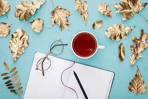 top view of tea in mug, glasses and notebook near golden foliage on blue background