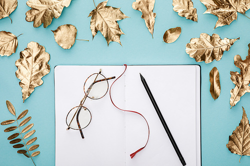 top view of autumnal golden foliage near blank notebook with glasses on blue background
