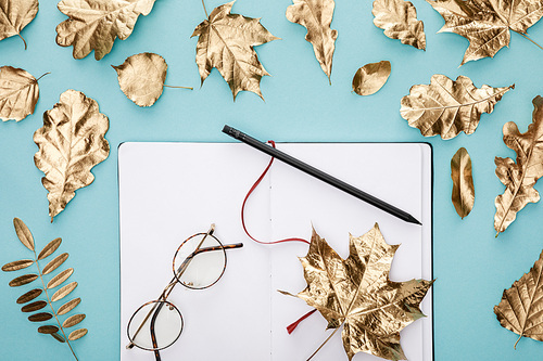 top view of autumnal golden leaves near blank notebook with glasses on blue background