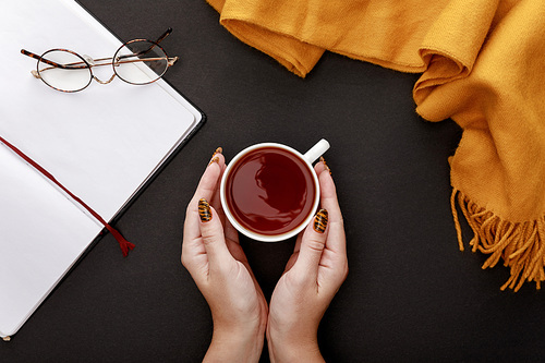 cropped view of woman holding tea in mug near glasses, scarf and blank notebook on black background