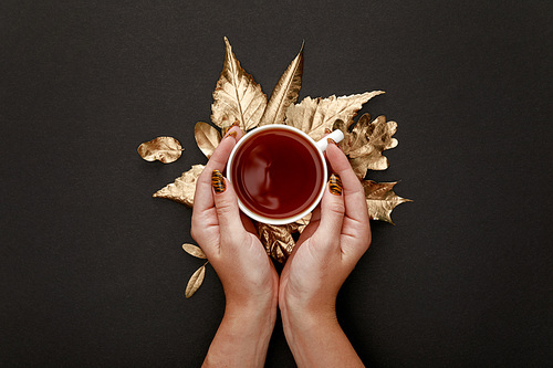 partial view of woman holding tea in mug near golden foliage on black background
