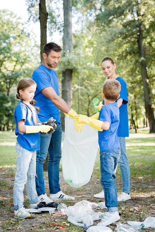 family of volunteers in rubber gloves collecting rubbish in recycled plastic bag in forest, ecology concept