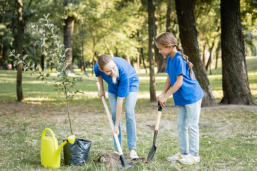 mother and daughter digging ground with shovels near watering can and seedling, ecology concept