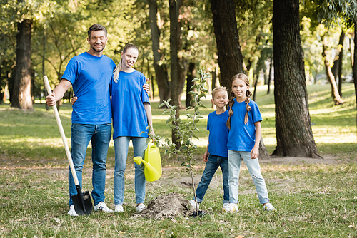 happy family of volunteers standing near planted young tree with shovel and watering can, ecology concept