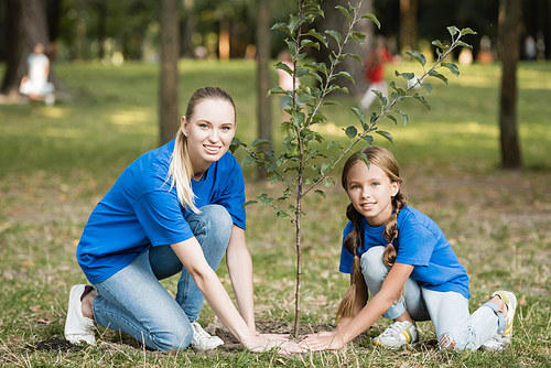 smiling mother and daughter  while planting young tree in park, ecology concept