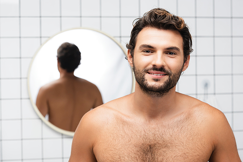 Shirtless man smiling at camera with mirror on blurred background in bathroom