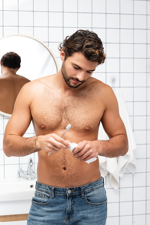 Shirtless man in jeans holding toothpaste and toothbrush in bathroom