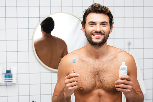 Cheerful shirtless man  while holding toothpaste and toothbrush in bathroom
