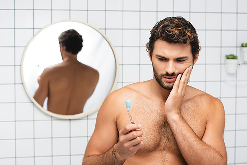 Shirtless man holding toothbrush while touching painful cheek in bathroom