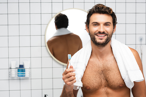 Smiling shirtless man with towel around neck holding toothbrush in bathroom