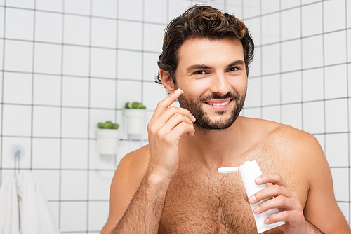 Shirtless man smiling while applying cosmetic cream in bathroom