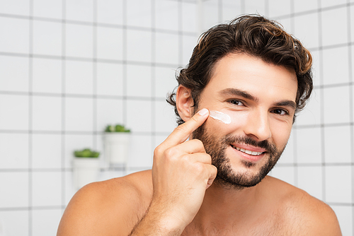 Smiling man applying cosmetic cream on face in bathroom