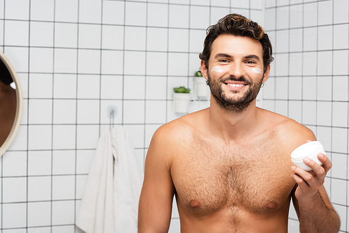 Smiling shirtless man holding jar with cosmetic cream in bathroom