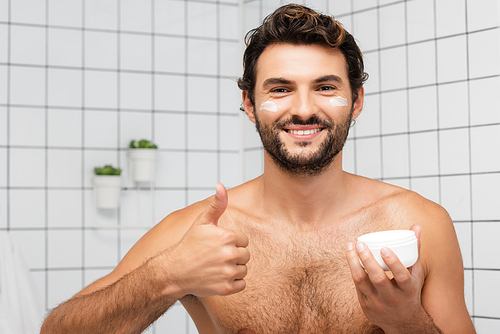 Smiling shirtless man showing like while holding jar with cosmetic cream in bathroom
