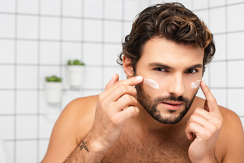 Bearded man applying cosmetic cream on face in bathroom