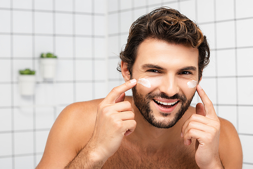 Cheerful shirtless man applying face cream and  in bathroom