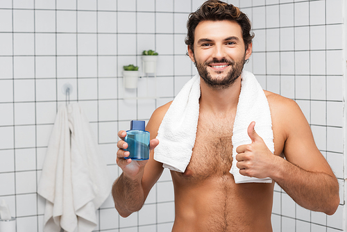 Smiling man with towel around neck showing thumb up and holding after shaving lotion in bathroom