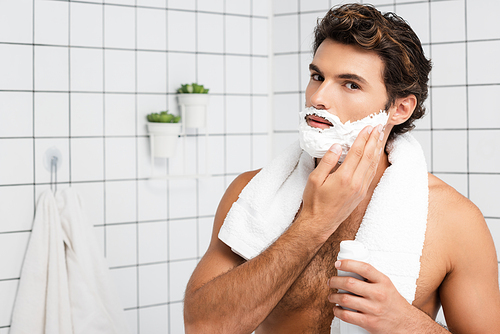 Muscular man with towel around neck applying shaving foam in bathroom