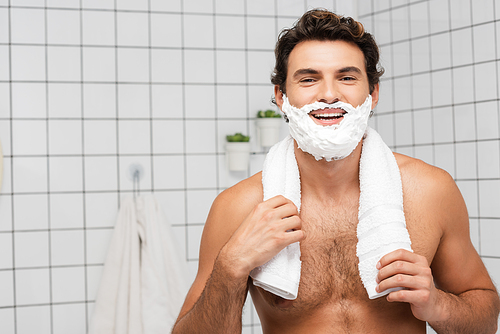 Cheerful shirtless man with shaving foam on face holding towel in bathroom