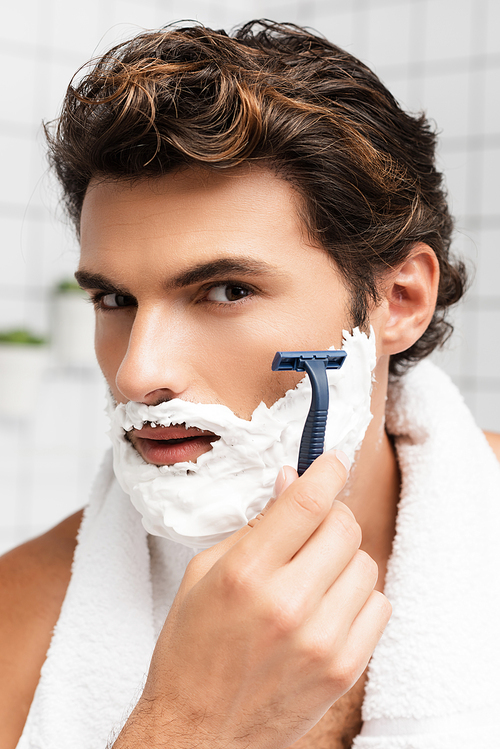 Brunette man with shaving foam on face shaving with razor in bathroom