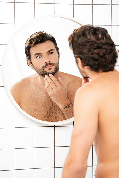 Shirtless man touching chin while looking at mirror in bathroom