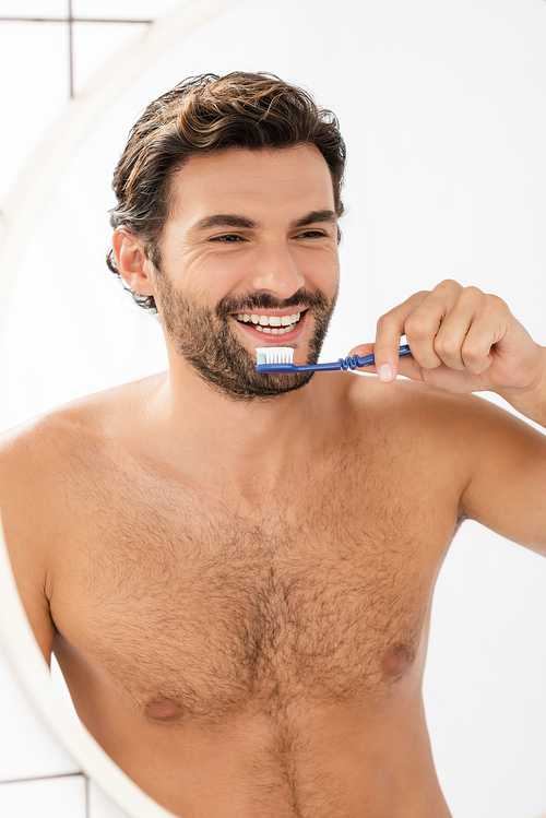 Shirtless man smiling at mirror while holding toothpaste and toothbrush in bathroom