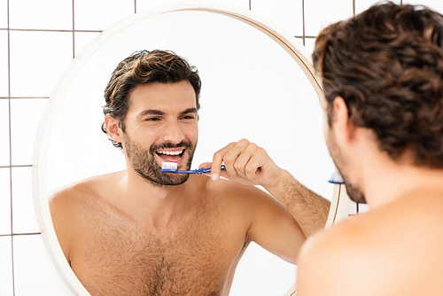 Smiling shirtless man reflecting in mirror while brushing teeth in bathroom