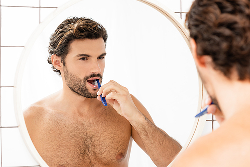 Shirtless man brushing teeth while looking at mirror in bathroom