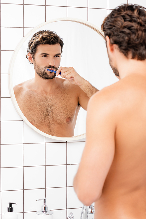 Barded and shirtless man looking at mirror while brushing teeth on blurred foreground in bathroom