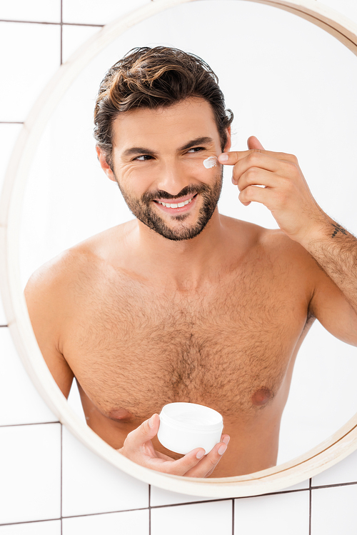 Cheerful man applying face cream while looking at mirror in bathroom