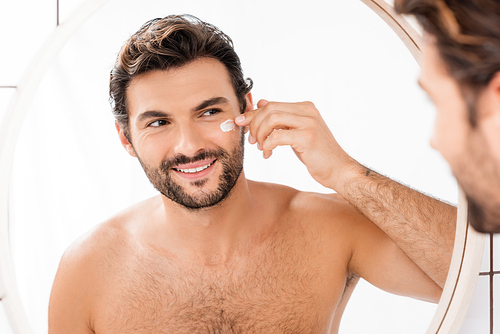 Smiling man looking at mirror while applying face cream on blurred foreground