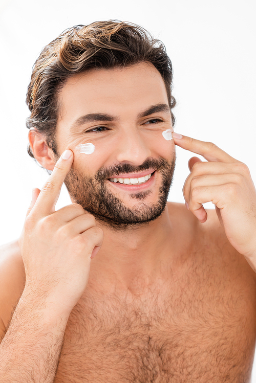 Smiling shirtless man applying face cream on cheeks isolated on grey