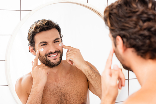 Positive man applying cosmetic cream on face near mirror on blurred foreground in bathroom