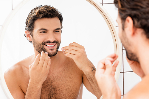Smiling shirtless man holding dental floss near mirror in bathroom