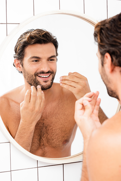 Shirtless man smiling while holding dental floss near mirror on blurred background