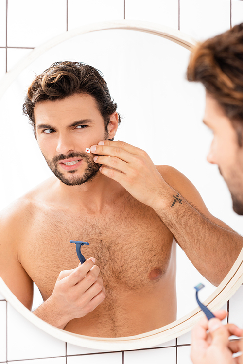Shirtless man touching cheek with wound and holding razor near mirror on blurred foreground