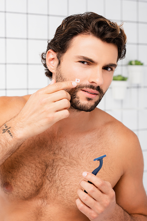 Shirtless man pointing at wound on cheek while holding razor in bathroom