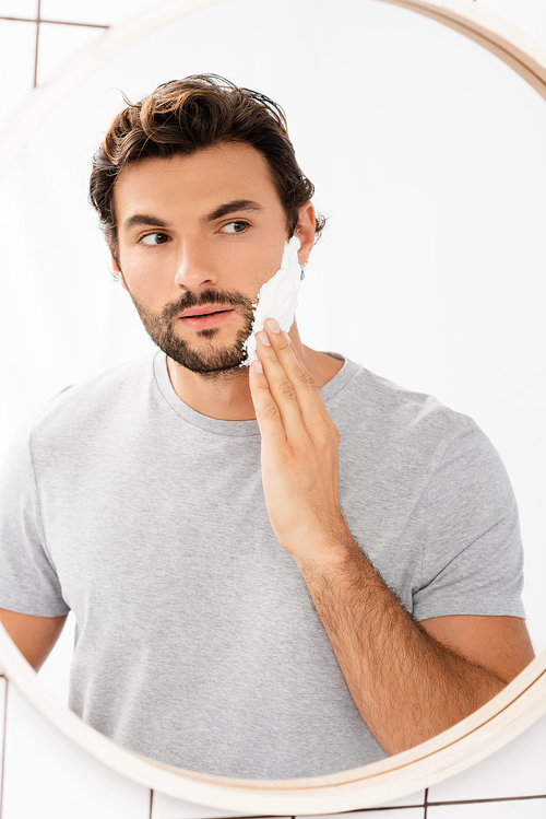 Bearded man looking at mirror while applying shaving foam in bathroom