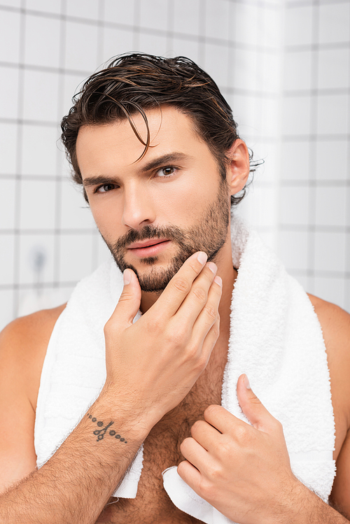 Shirtless man with wet hair and towel around neck touching chin in bathroom
