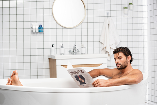 Bearded man with business lettering on newspaper taking bath at home