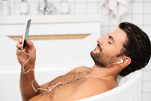 Young man in earphones taking bath and using smartphone