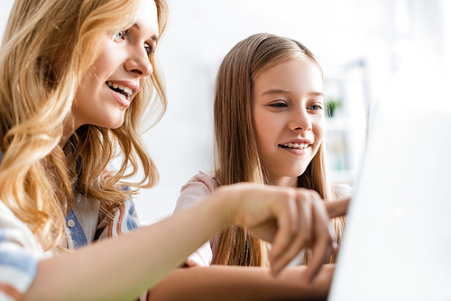 Selective focus of mother pointing with finger at laptop near smiling child at home