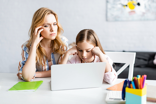 Selective focus of pensive mother and kid looking at laptop during online education at home