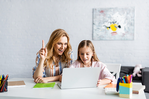 Selective focus of smiling woman having idea while using laptop near kid and stationery on table
